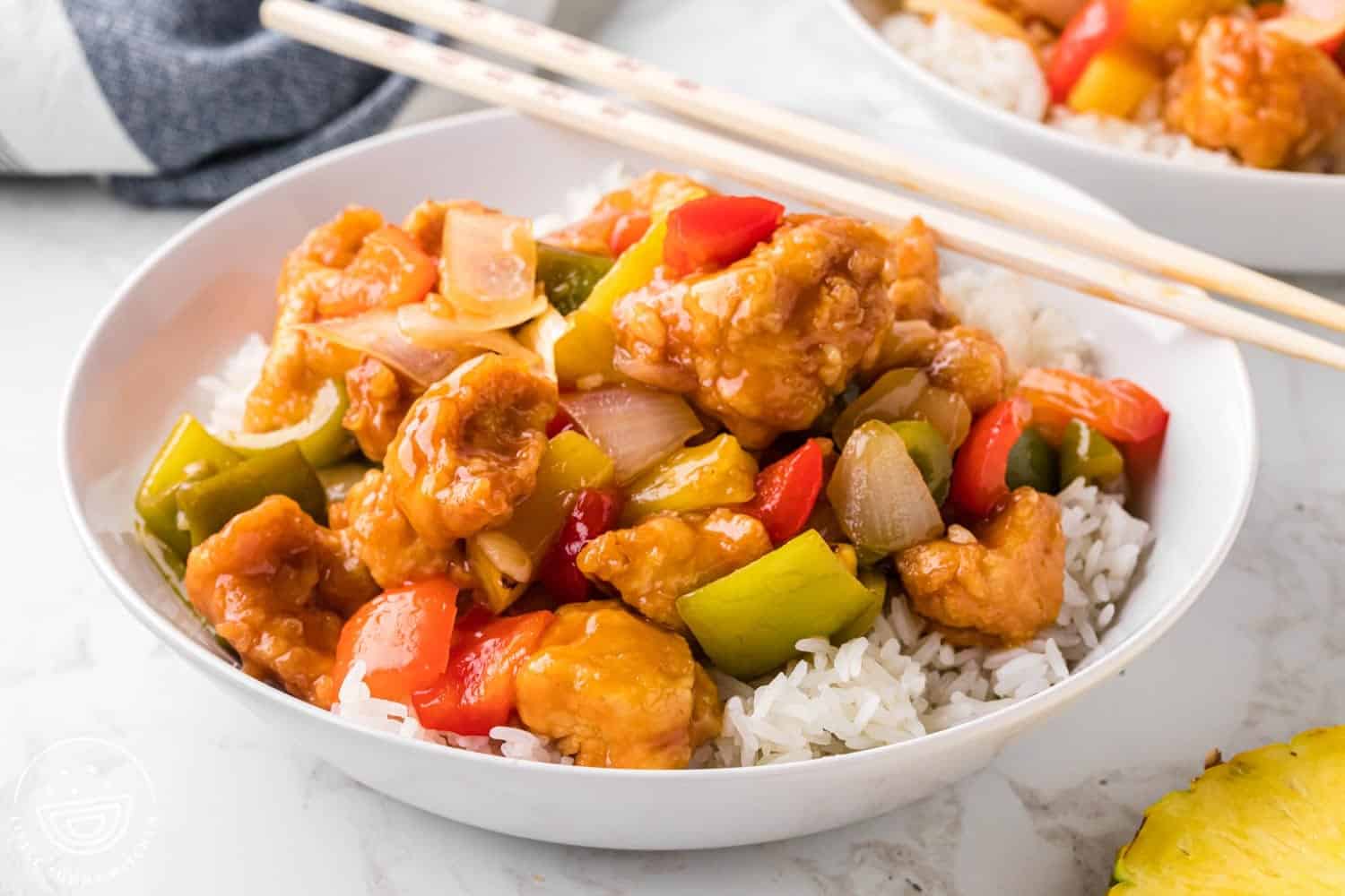 Horizontal image of sweet and sour chicken served over rice in a white bowl, with chop sticks on the side.