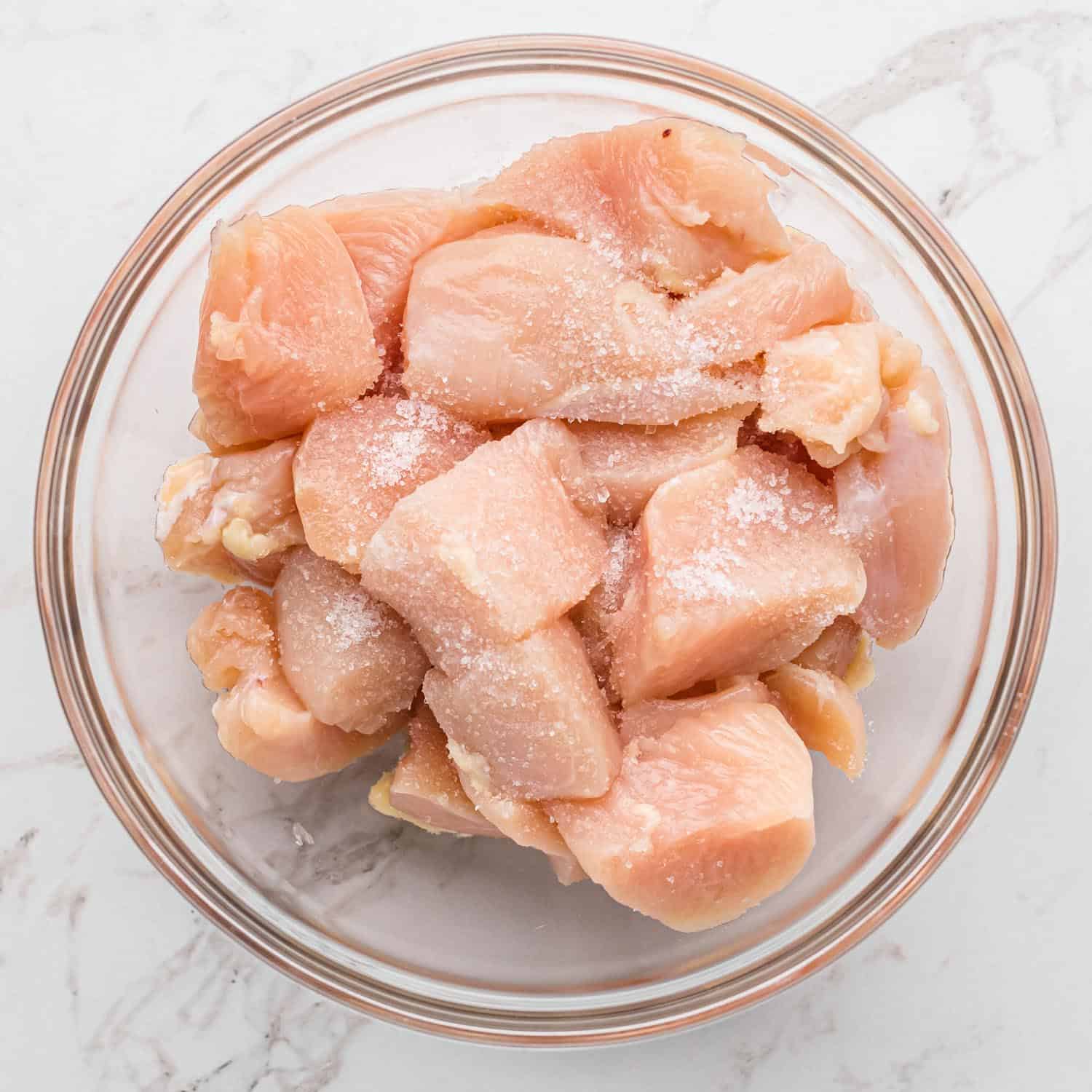 Cubed chicken in a large glass bowl, seasoned with salt.