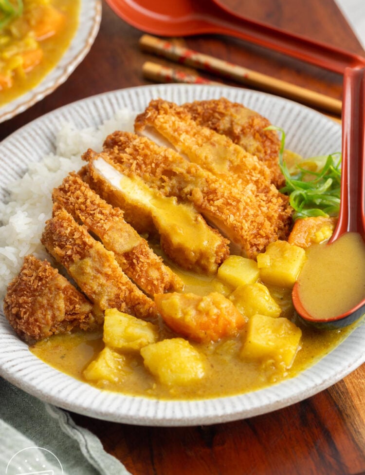 A bowl of white rice, sliced chicken katsu and curry with a japanese spoon and chopsticks.