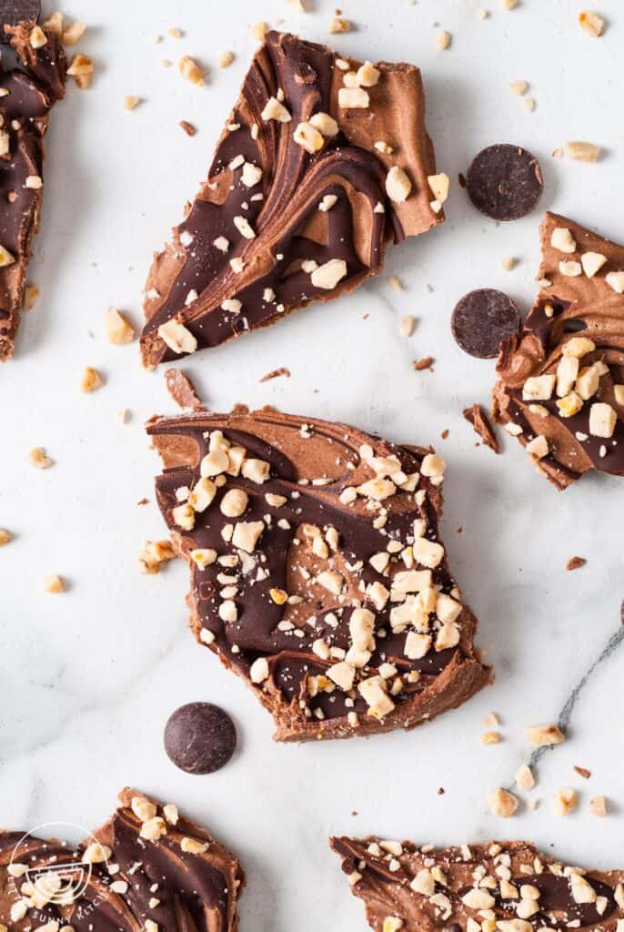 uneven pieces of frozen chocolate bark on a marble counter. The yogurt bark is topped with melted chocolate drizzle and chopped toffee bits.