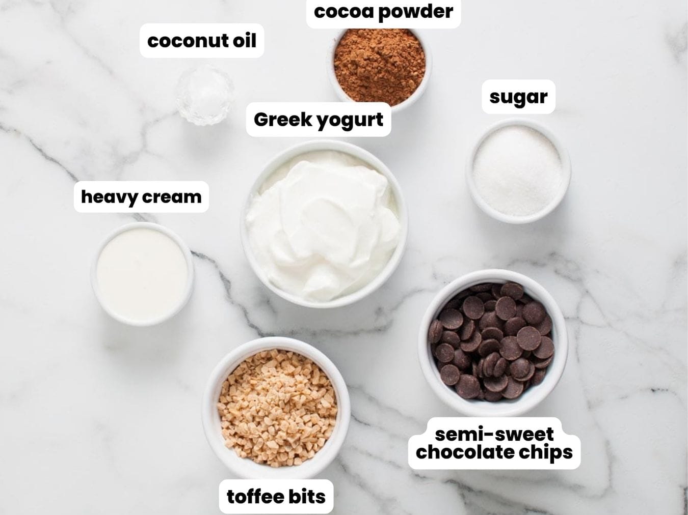 The ingredients needed for making chocolate yogurt bark, all in separate bowls, including yogurt, cocoa powder, toffee bits, coconut oil, chocolate chips, and sugar.