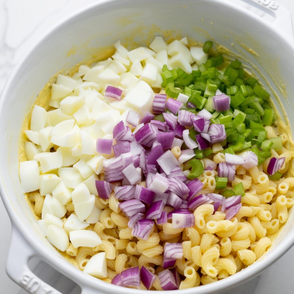 chopped egg whites, red onion, and celery added to a bowl with cooked macaroni and dressing.