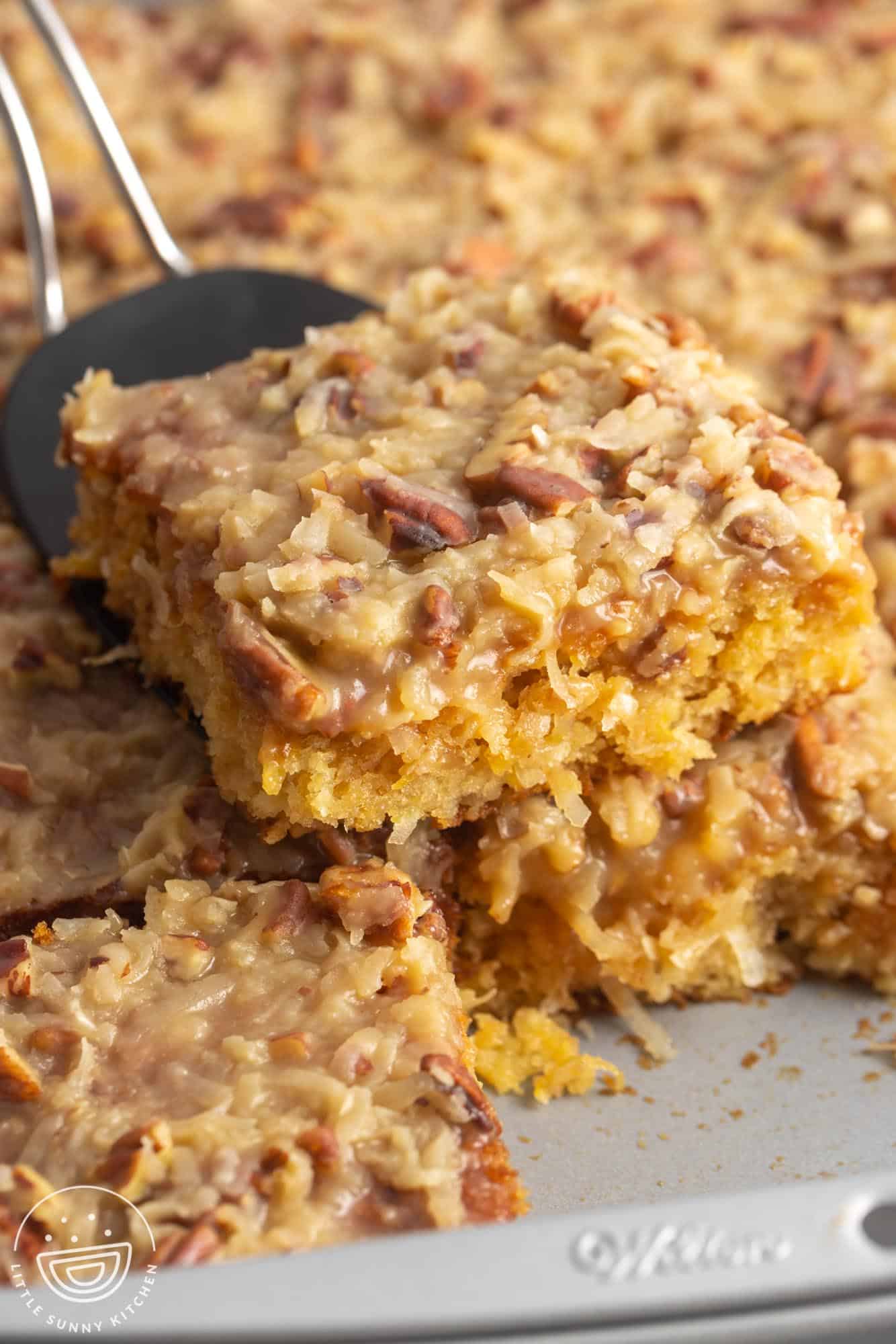 a spatula lifting a slice of Do Nothing Cake from a pan to show the tender pineapple cake topped with coconut pecan frosting.