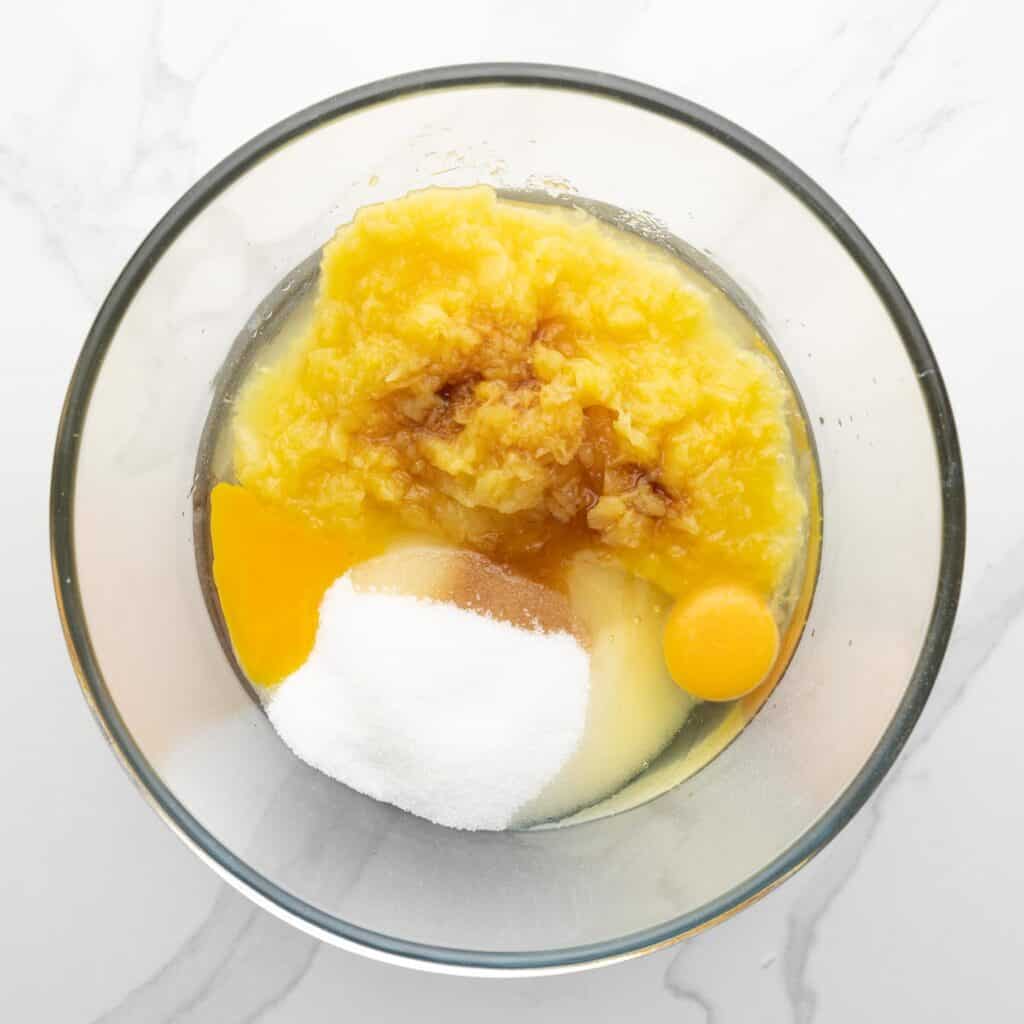 a glass mixing bowl holding crushed pineapple, eggs, sugar, and vanilla extract.