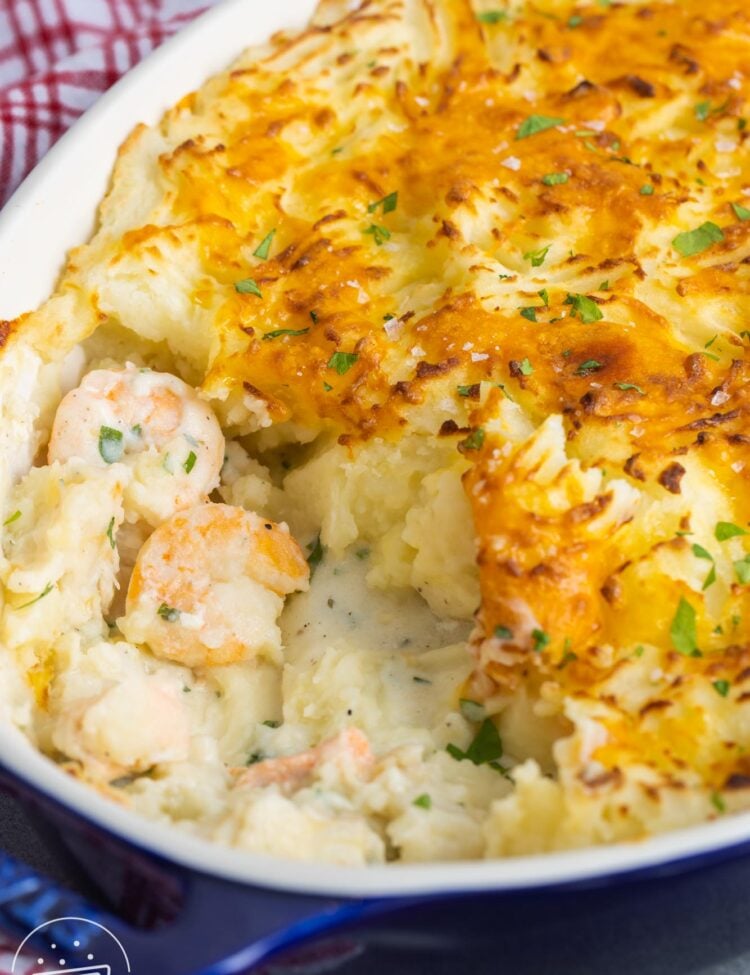 fish pie in a blue casserole dish. Some has been served, showing the creamy fish and shrimp stew under the crispy mashed potatoes.