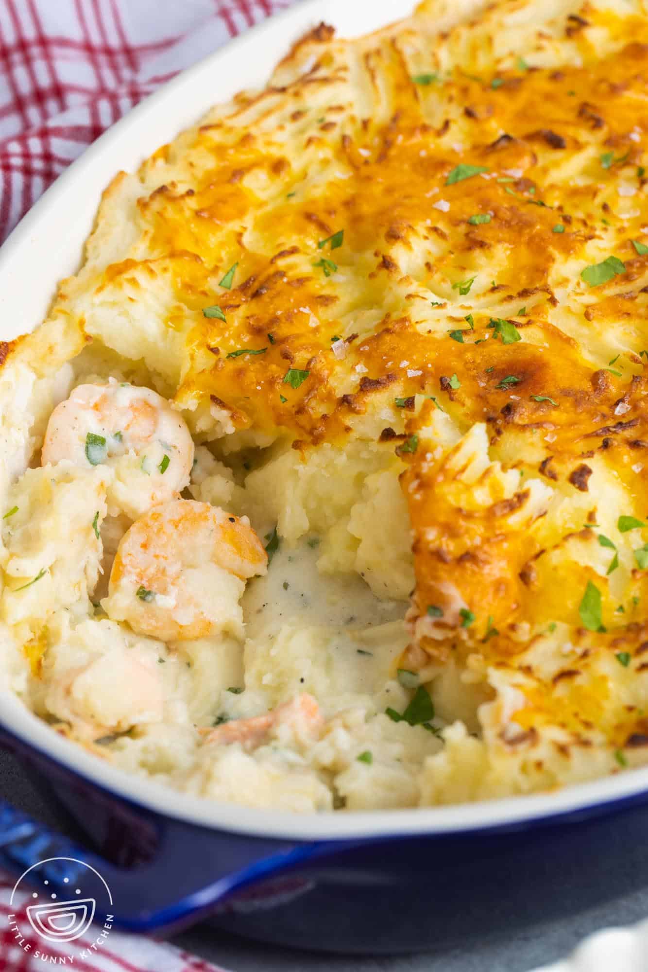 fish pie in a blue casserole dish. Some has been served, showing the creamy fish and shrimp stew under the crispy mashed potatoes.