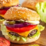 a cheeseburger on a sesame seed roll topped with thick slices of red onion and tomato, pickle chips, and sauce.