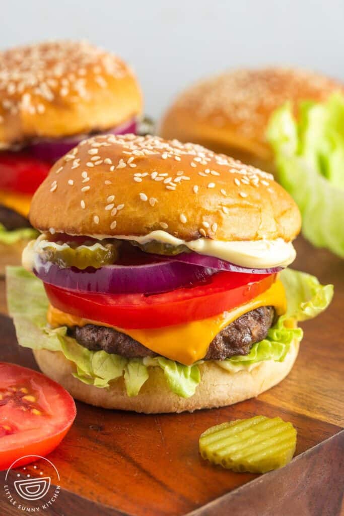 a cheeseburger on a sesame seed roll topped with thick slices of red onion and tomato, pickle chips, and sauce.