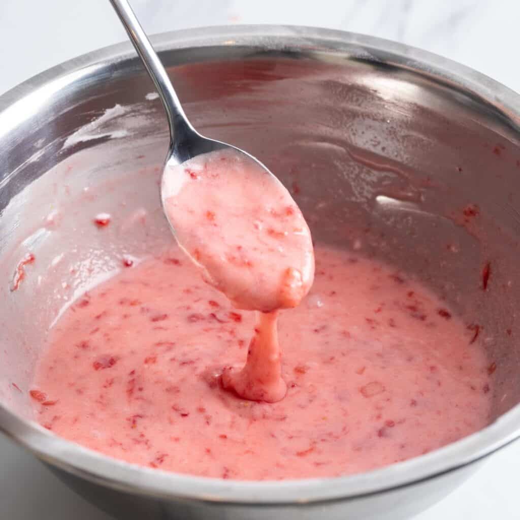 strawberry icing in a metal mixing bowl, drizzled from a spoon.