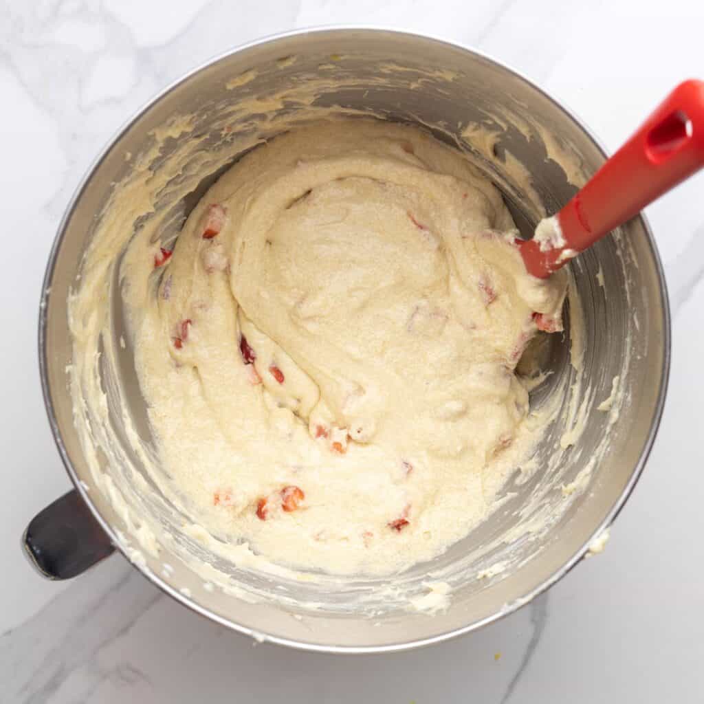 A bowl of vanilla pound cake batter with diced strawberries stirred in with a red spatula.