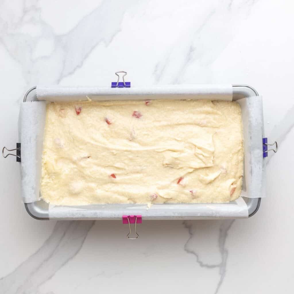 a loaf pan lined with parchment paper. Binder clips are holding the paper on to each side. Inside the pan is strawberry pound cake batter.