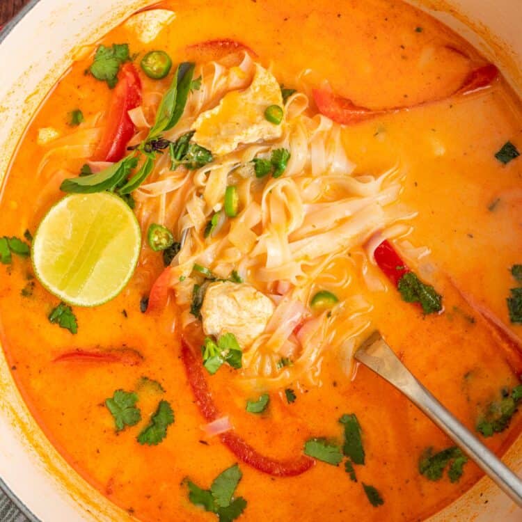 Overhead view of Thai curry soup with chicken, noodles, and red peppers, garnished with fresh herbs and lime wedges.