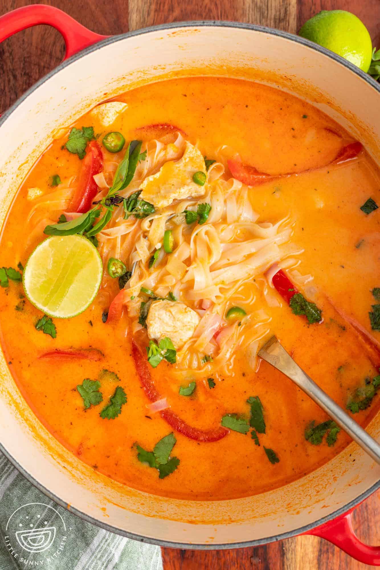 Overhead view of Thai curry soup with chicken, noodles, and red peppers, garnished with fresh herbs and lime wedges.