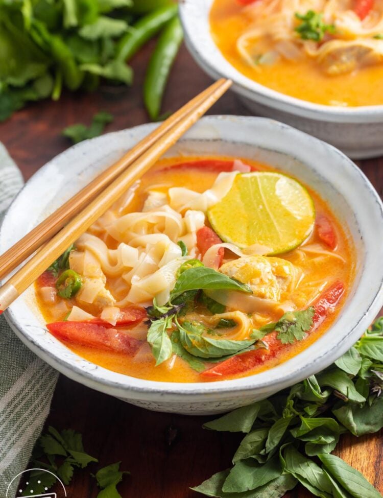 A bowl of thai curry soup with noodles and chicken. Chopsticks rest on the side of the bowl, and it's garnished with fresh cilantro and thai basil.