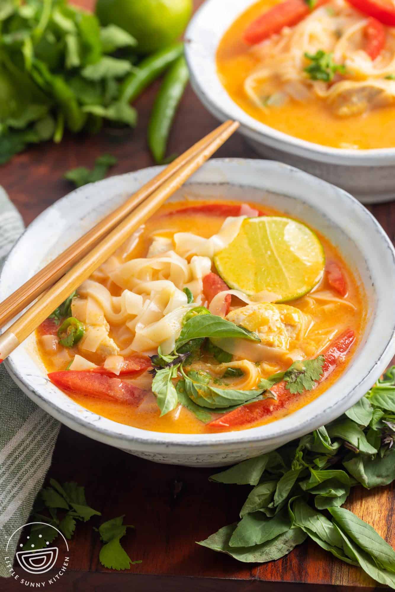 A bowl of thai curry soup with noodles and chicken. Chopsticks rest on the side of the bowl, and it's garnished with fresh cilantro and thai basil.