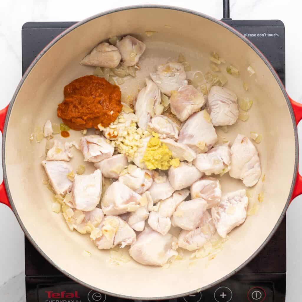 Chunks of briefly cooked chicken in a dutch oven with red curry paste, garlic, and ginger