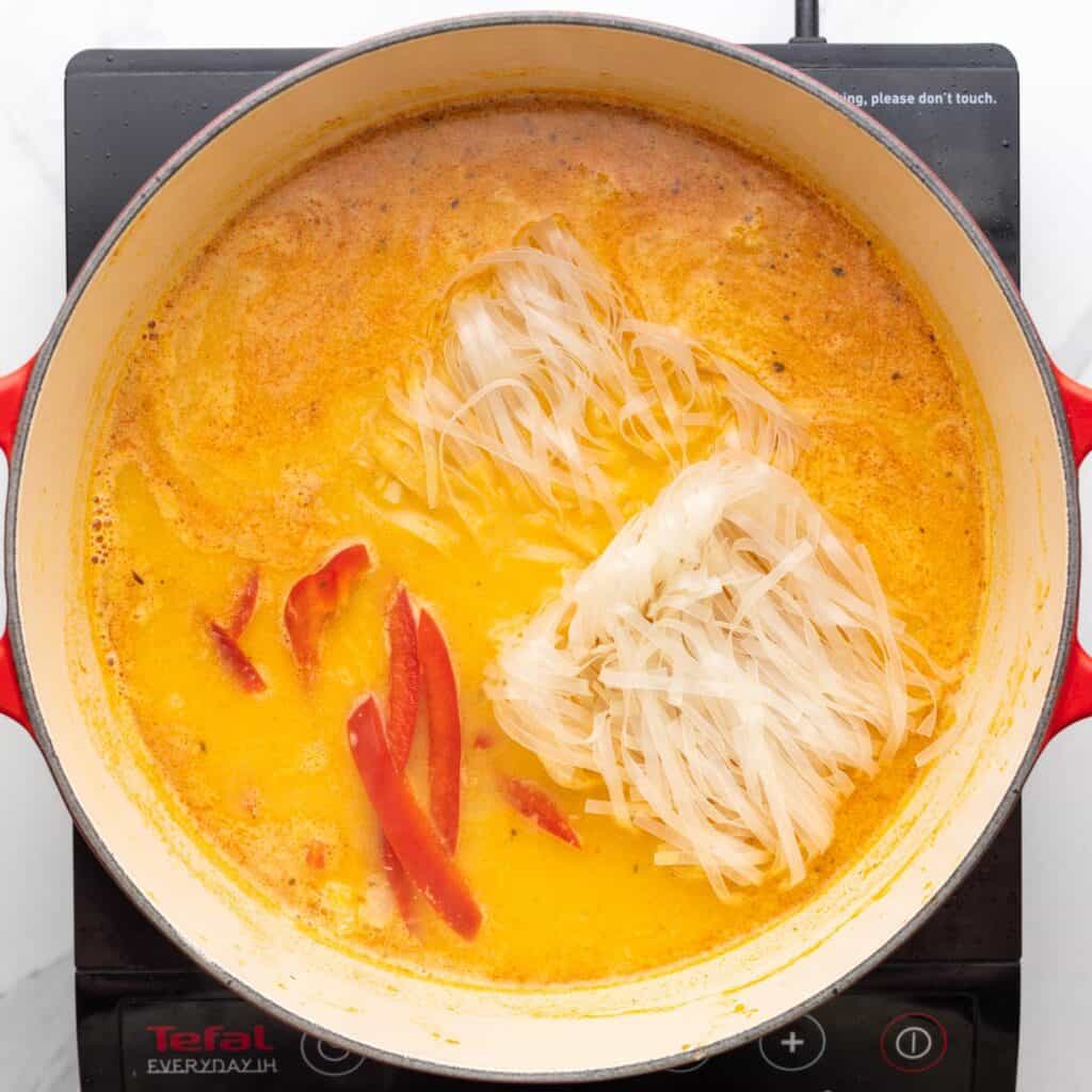 Dry rice noodles and sliced red peppers added to a pot of curry soup.