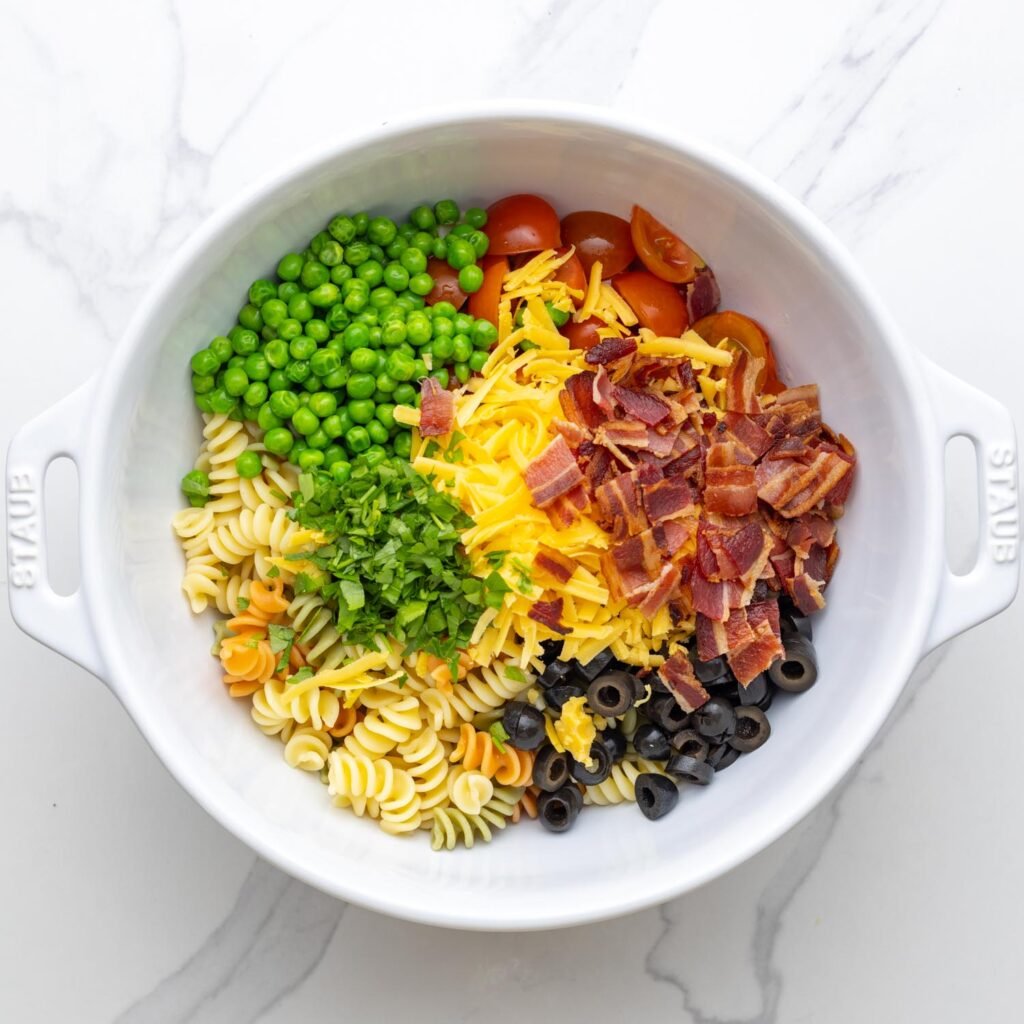 a large white bowl of pasta topped with shredded cheese, diced bacon, sliced olives, tomatoes, peas, and parsley.