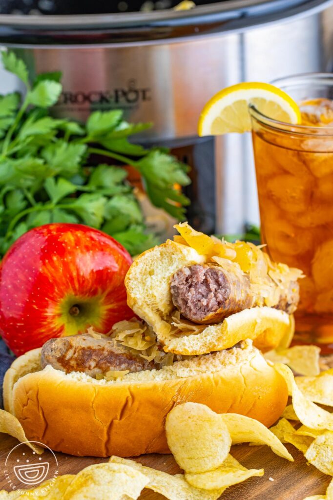 Two sausages in rolls, topped with sauerkraut. There are potato chips next to them. In the background is an apple, a glass of iced tea, and a crock pot.