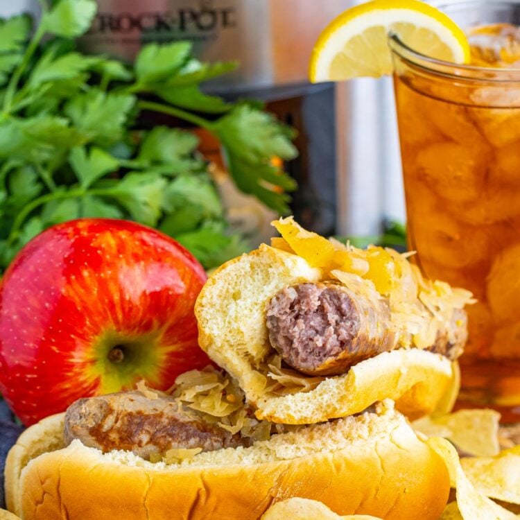 Two sausages in rolls, topped with sauerkraut. There are potato chips next to them. In the background is an apple, a glass of iced tea, and a crock pot.