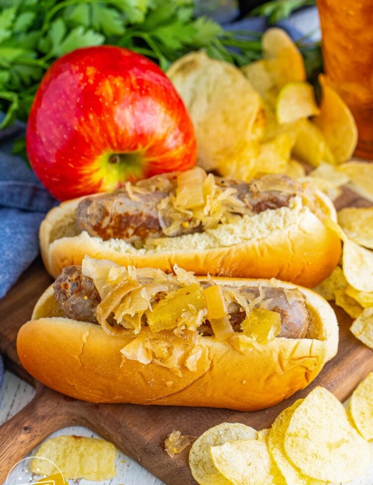 overhead view of two bratwurst sandwiches topped with sauerkraut. The sausages are on a wooden cutting board next to a red apple and potato chips.