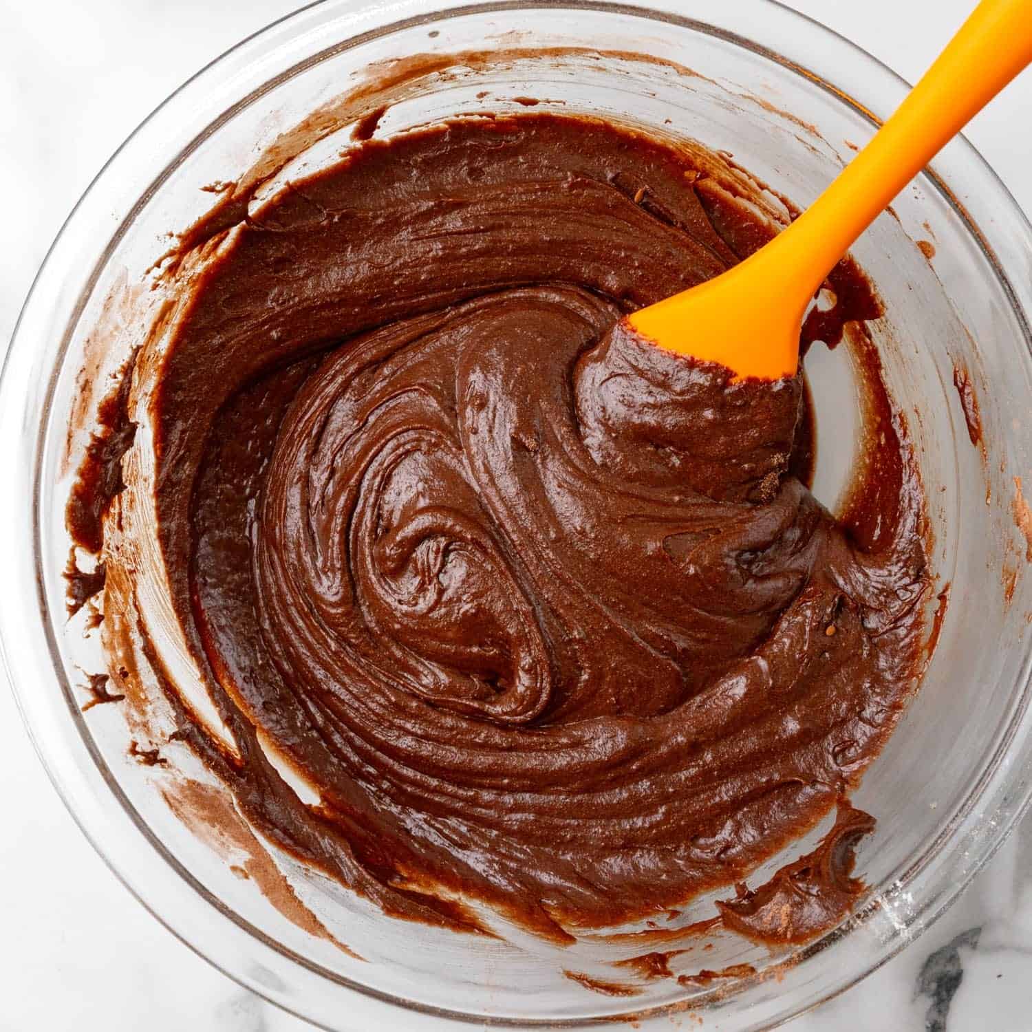 Brownie batter stirred with an orange spatula in a large glass mixing bowl.