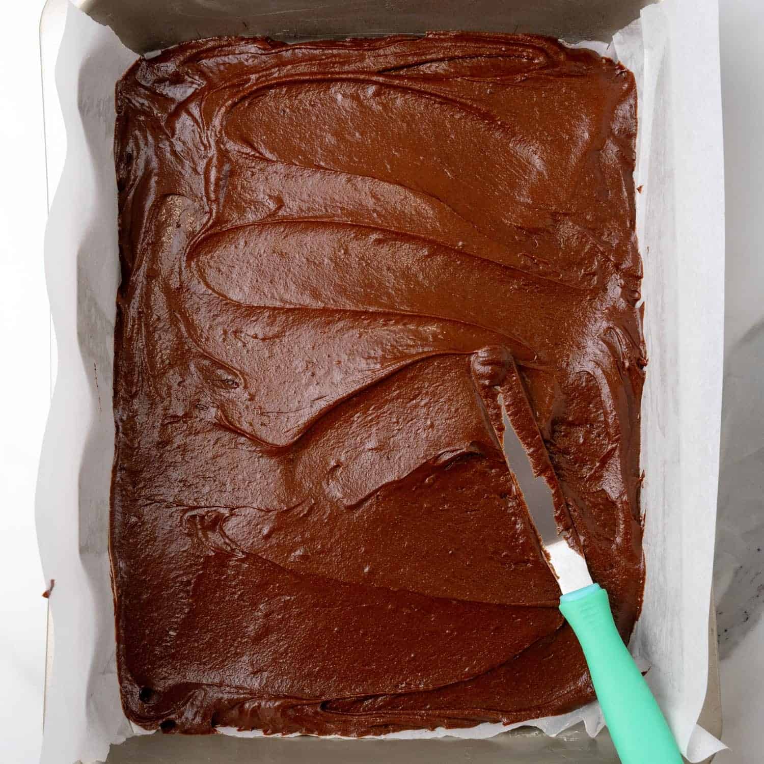 An offset spatula spreading brownie batter into a metal pan that has been lined with parchment paper.