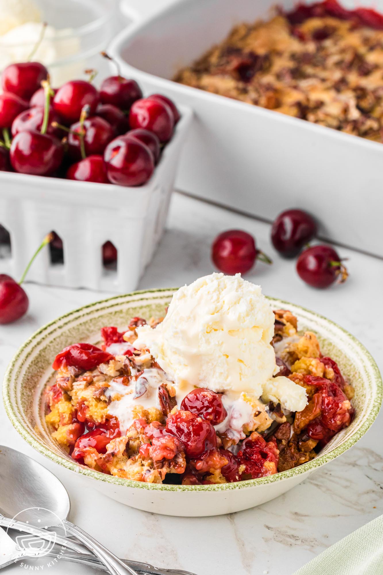 a bowl of cherry dump cake topped with vanilla ice cream, in front of a box of fresh cherries and the full pan of dump cake.