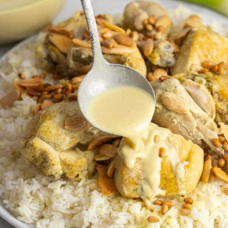 Pouring yogurt sauce on a piece of chicken, on chicken mansaf served on a platter with toasted nuts and rice.