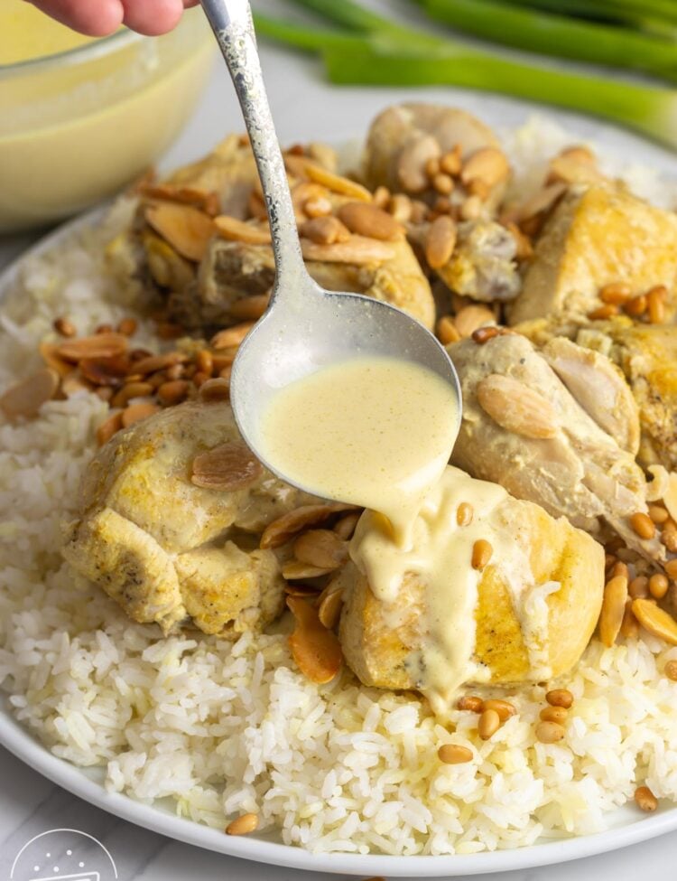 Pouring yogurt sauce on a piece of chicken, on chicken mansaf served on a platter with toasted nuts and rice.
