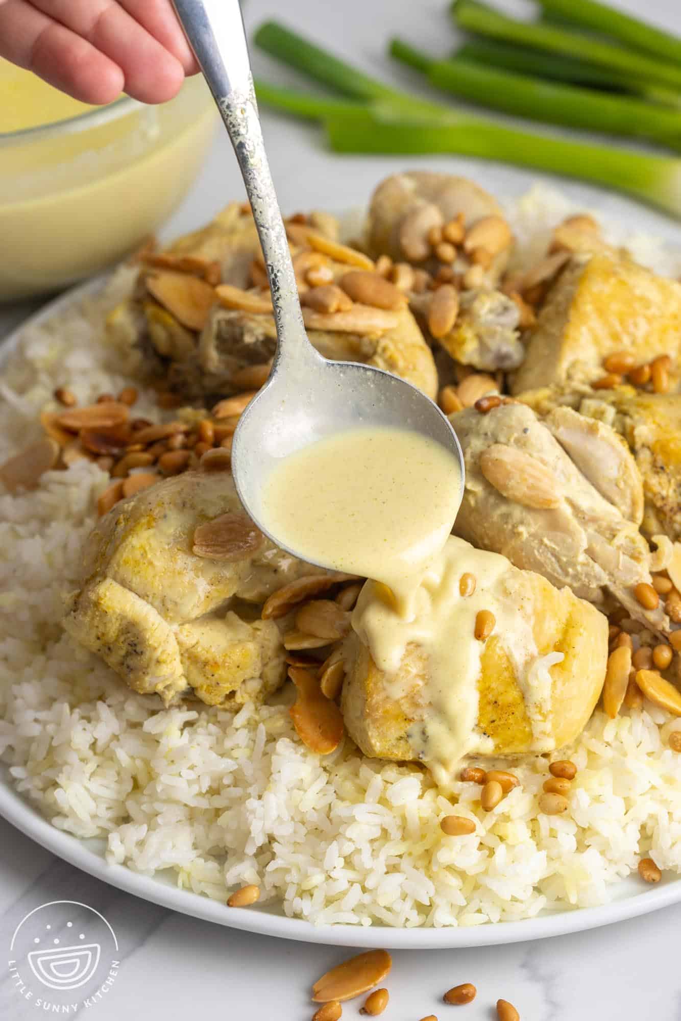 Pouring yogurt sauce on a piece of chicken, on chicken mansaf served on a platter with toasted nuts and rice.