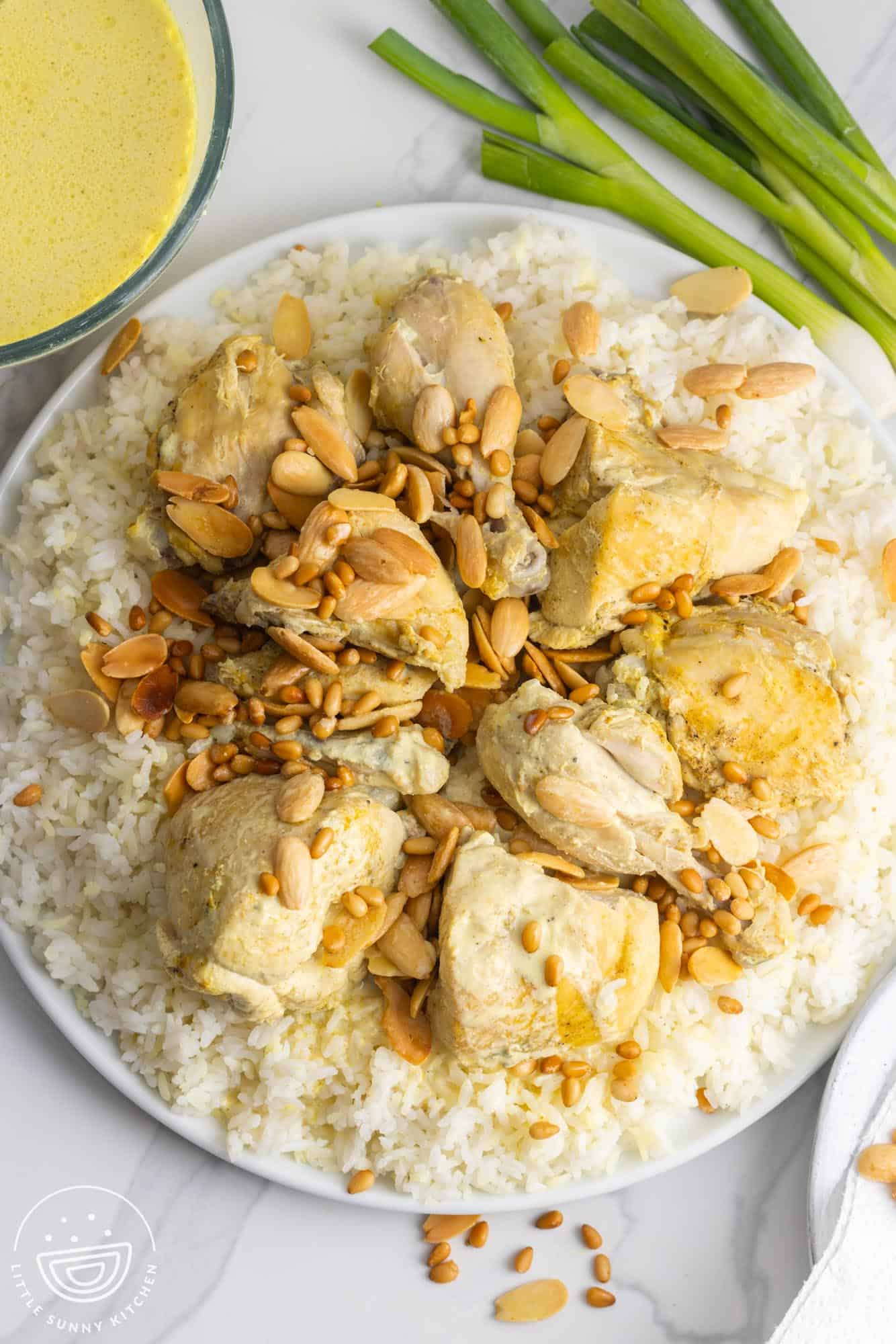 Overhead shot of a platter with chicken mansaf over a bed of rice, with yogurt sauce on the side, and green onions.