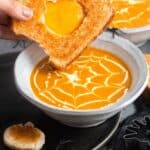 a hand dipping a grilled cheese sandwich into halloween pumpkin soup