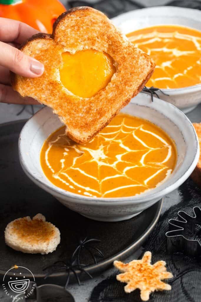 a hand dipping a grilled cheese sandwich into halloween pumpkin soup