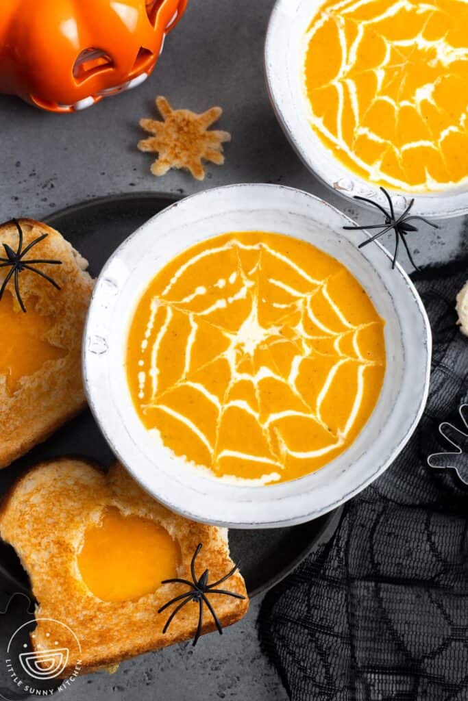 bowls of pumpkin soup with cream drizzles made to look like spider webs. Soup is served with grilled cheese sandwiches with fun halloween shape cutouts. Plastic spiders decorate the table.