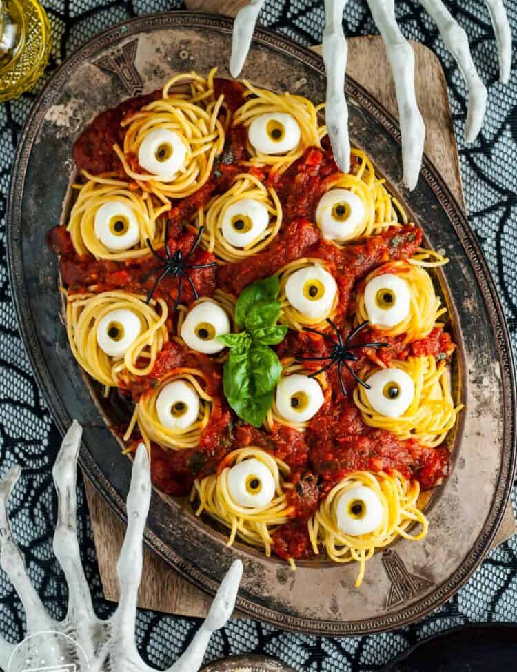 A platter of halloween spaghetti with mozzarella eyes on a spooky table with skeleton hand decorations.