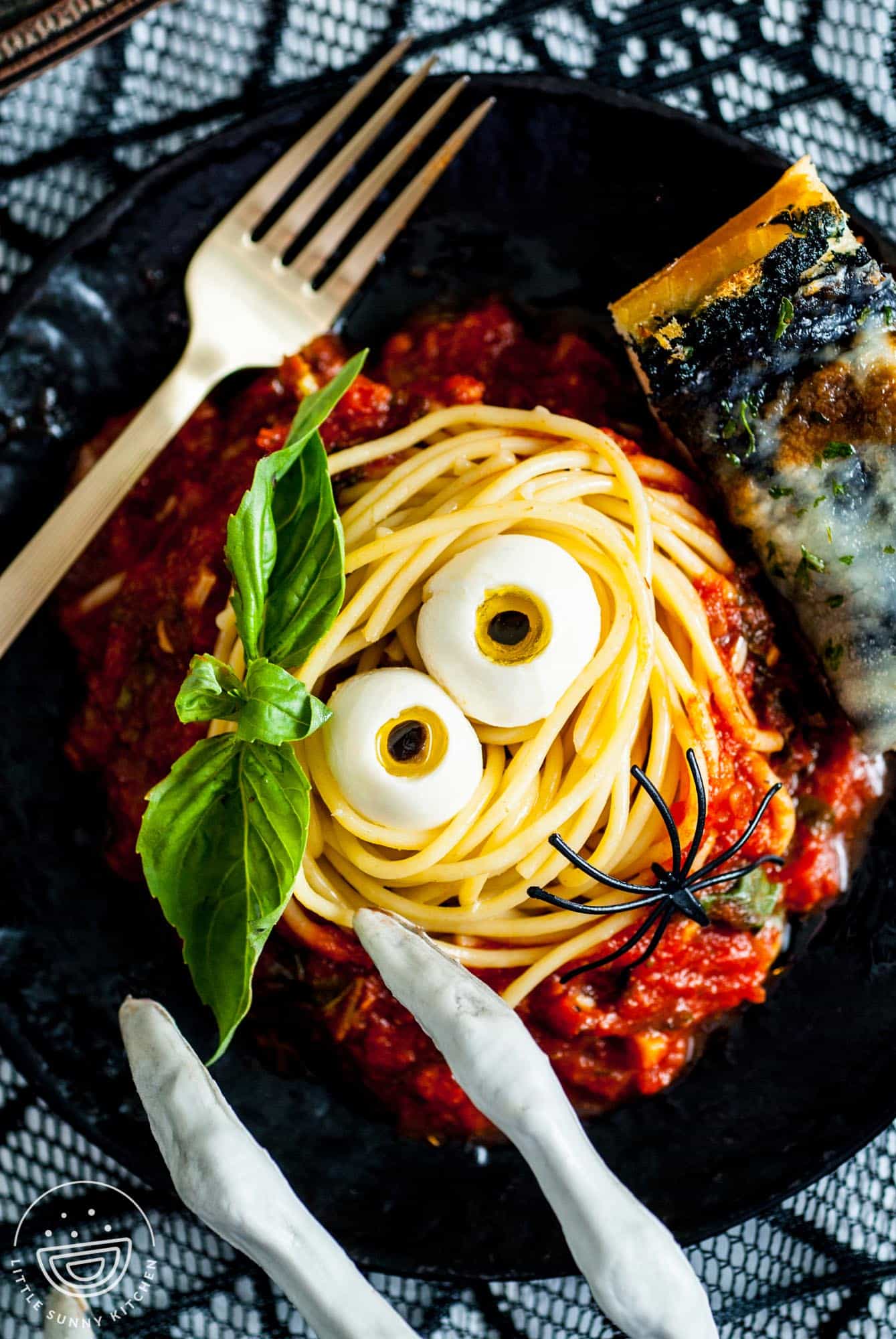 a black plate of Halloween Spaghetti. A nest of pasta is around two edible eyeballs, surrounded by pasta sauce, garnished with basil and a piece of black garlic bread.