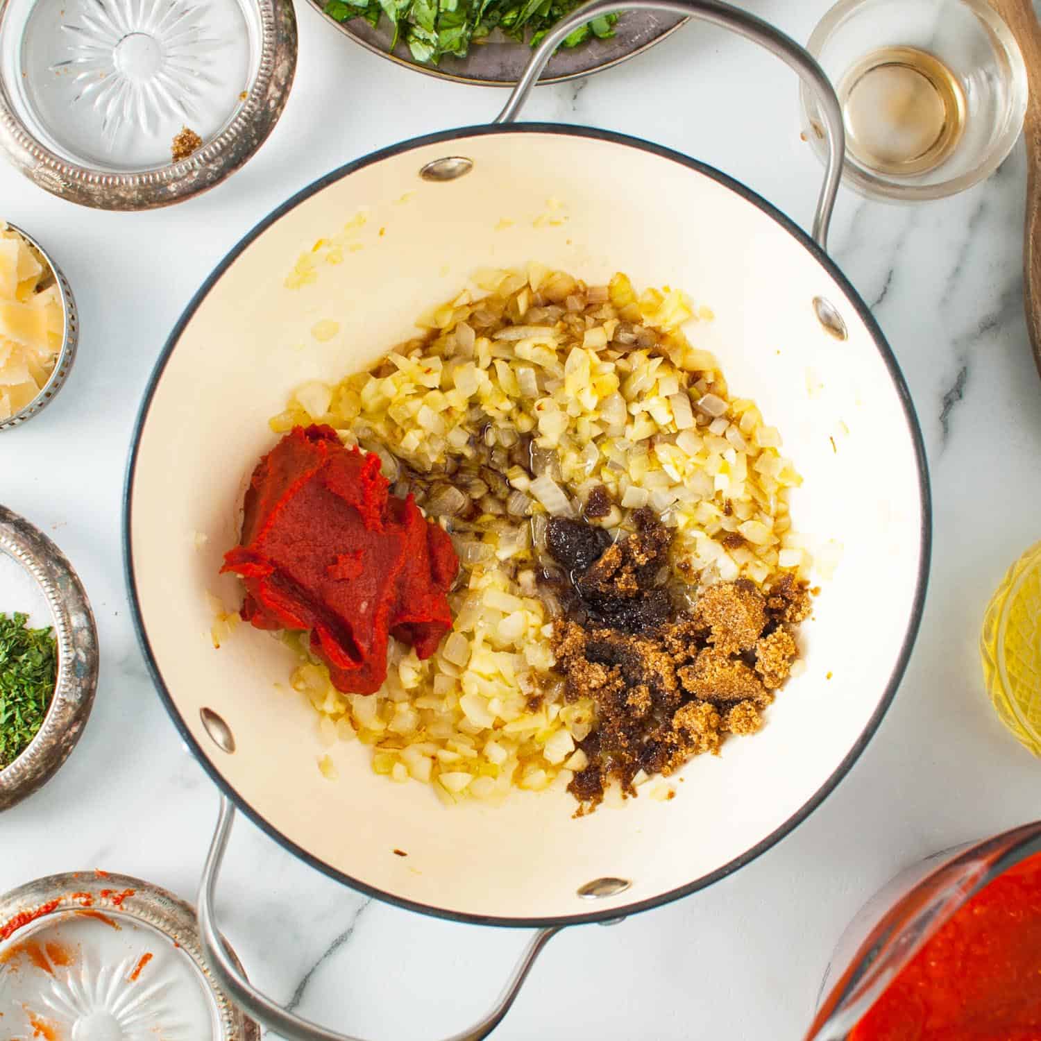 tomato paste and brown sugar added to softened onions in a dutch oven. Surrounding the pot are ornate bowls with other ingredients.