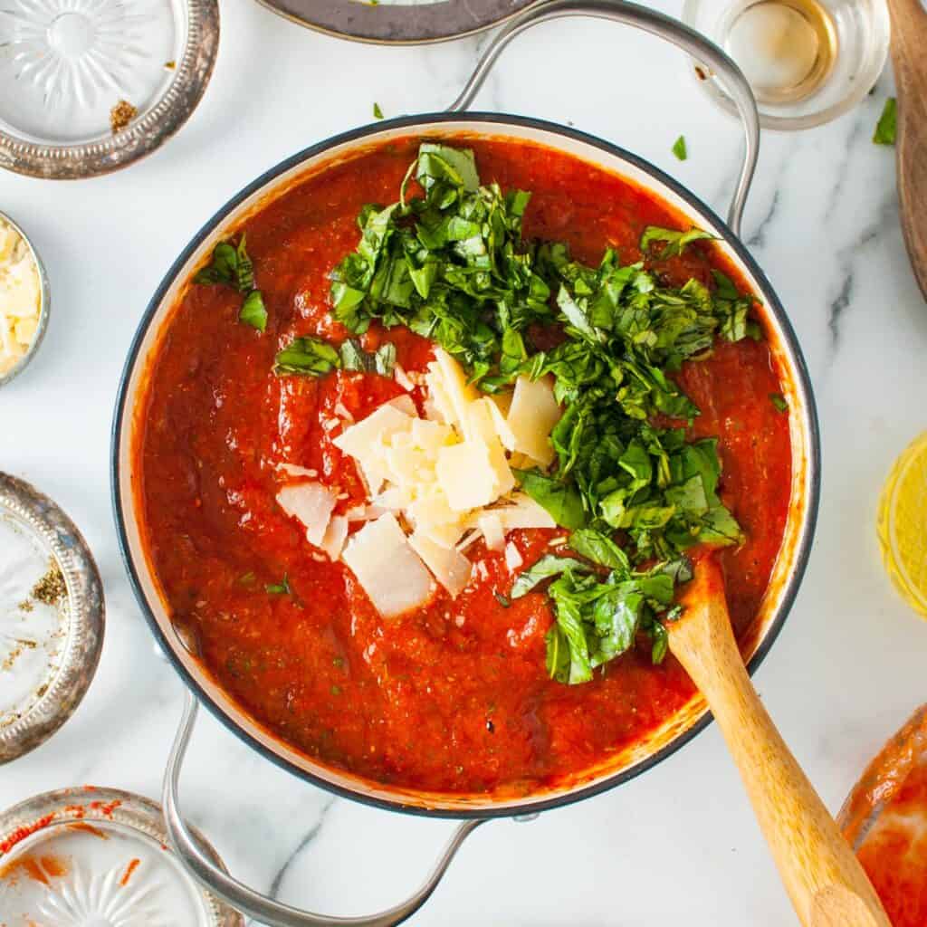 chopped basil and coarsely shredded parmesan cheese added to a pot of tomato sauce that is stirred with a wooden spoon.