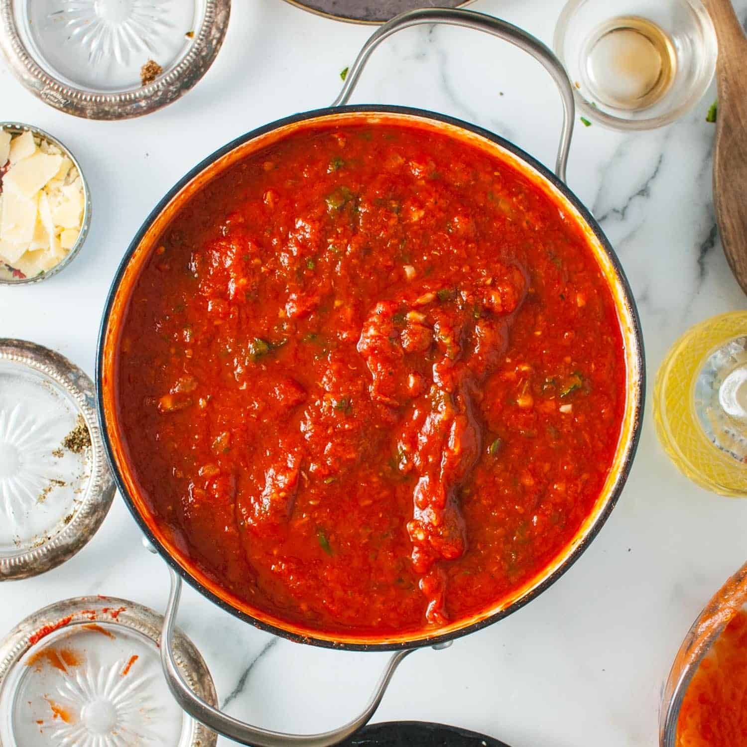 Homemade tomato sauce for halloween spaghetti is cooked in a dutch oven and sitting on a marble counter. Surrounding the pot are empty ingredient bowls.