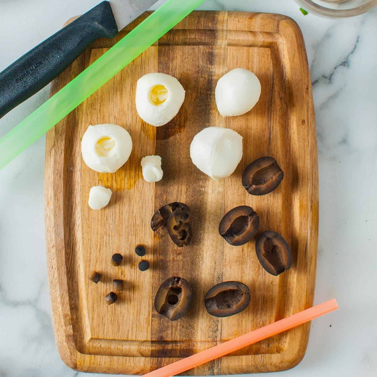 Mozzarella balls and black olives are on a small wooden cutting board. Straws have been used to cut small black circles from the olives and wider holes from the cheese.