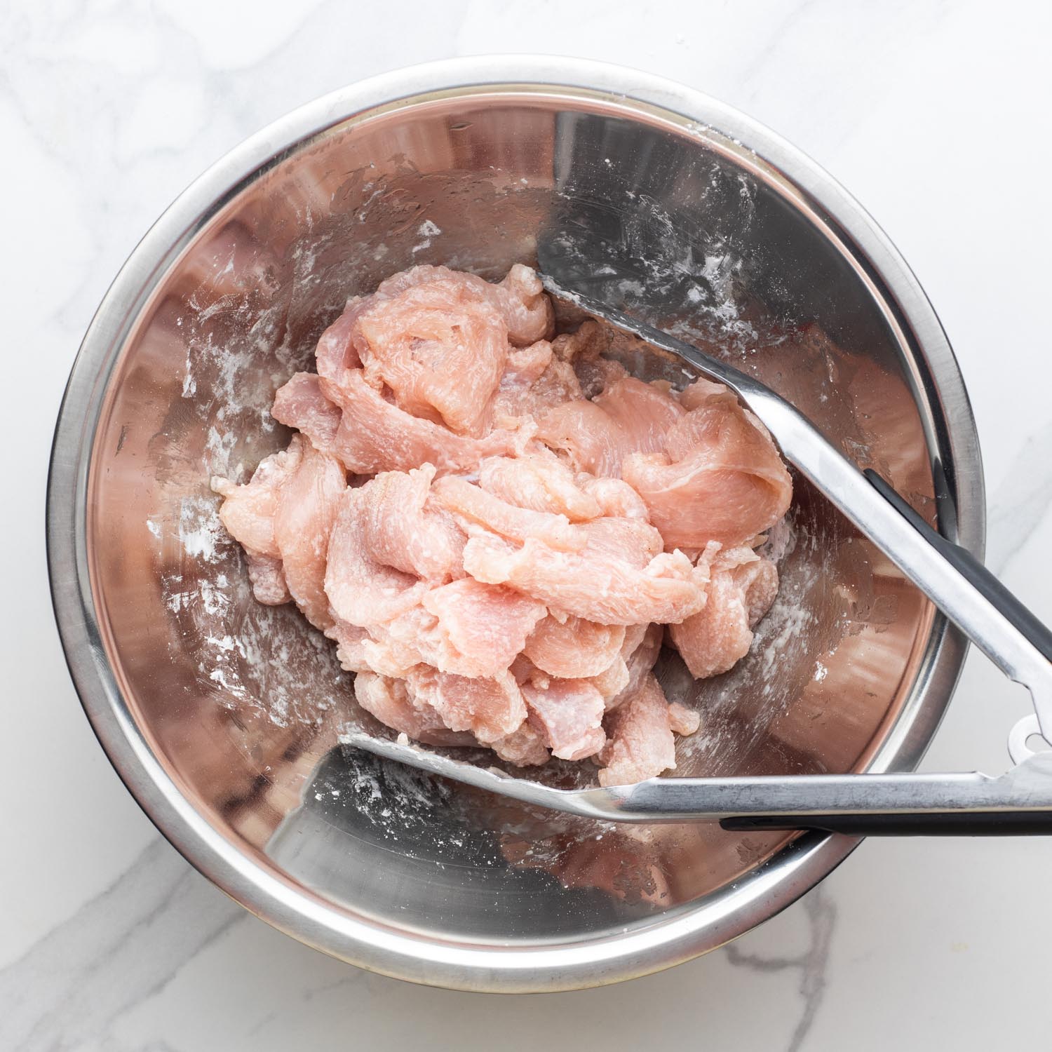 thin slices of chicken breast tossed with cornstarch and seasonings in a small bowl with tongs.