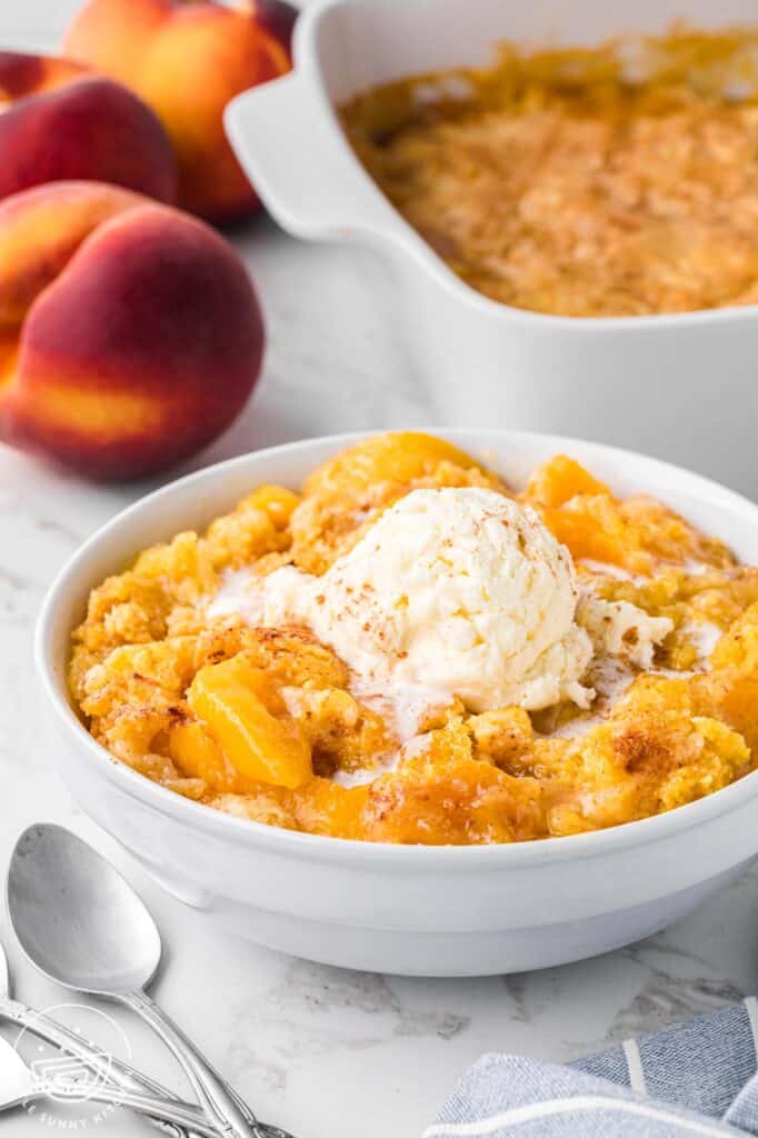 a bowl of peach dump cake with vanilla ice cream in front of a pan of the dessert and some fresh peaches.