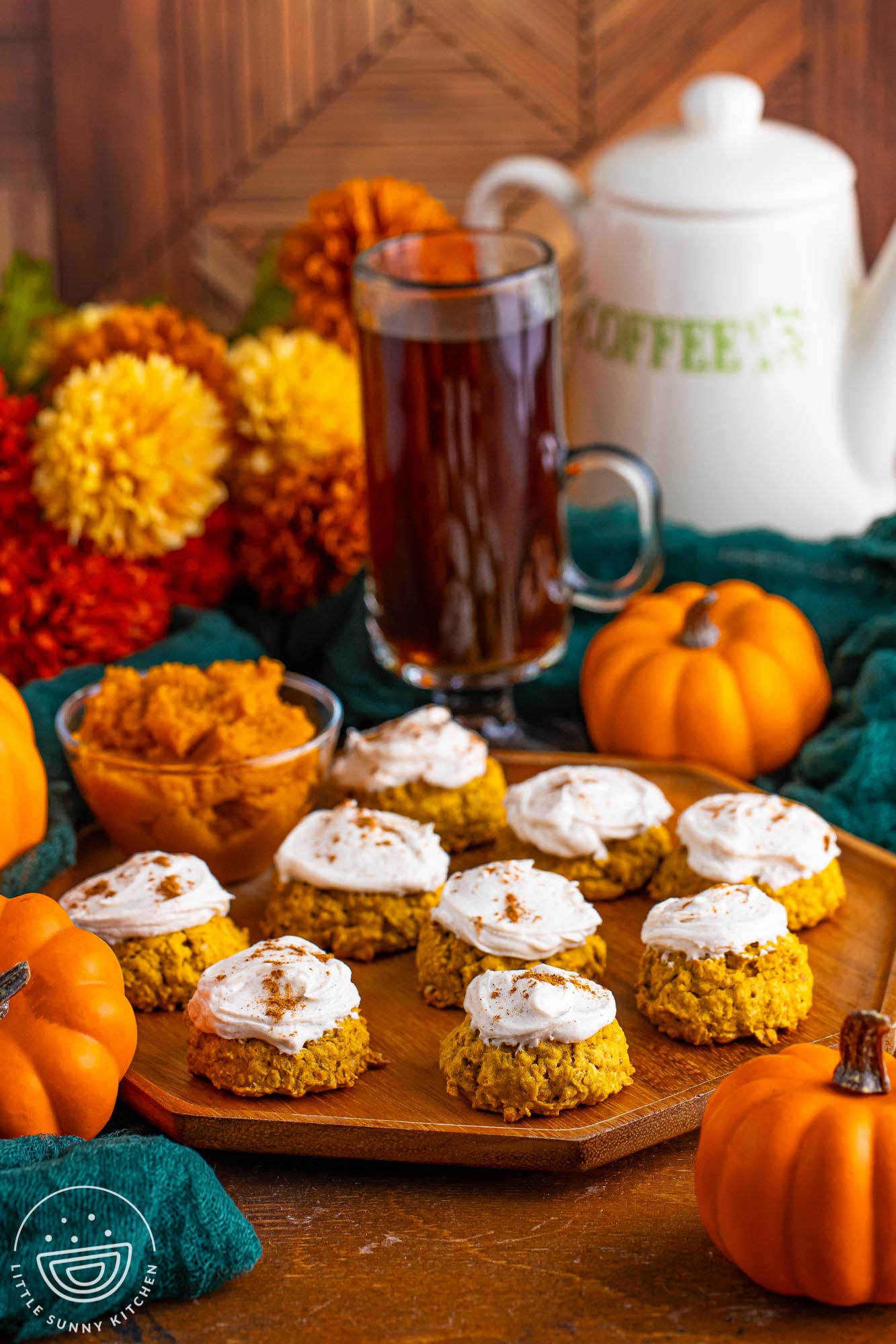 a tray of pumpkin oatmeal cookies. In the background are small pumpkins, fall mums, and a tall coffee in a glass mug.