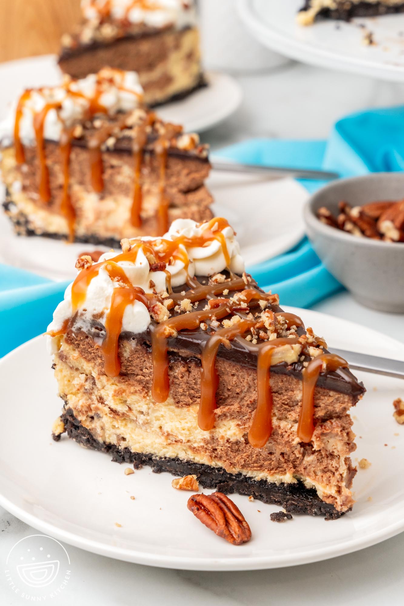 a white dessert plate holding a slice of turtle cheesecake that has been drizzled with caramel sauce and pecans.