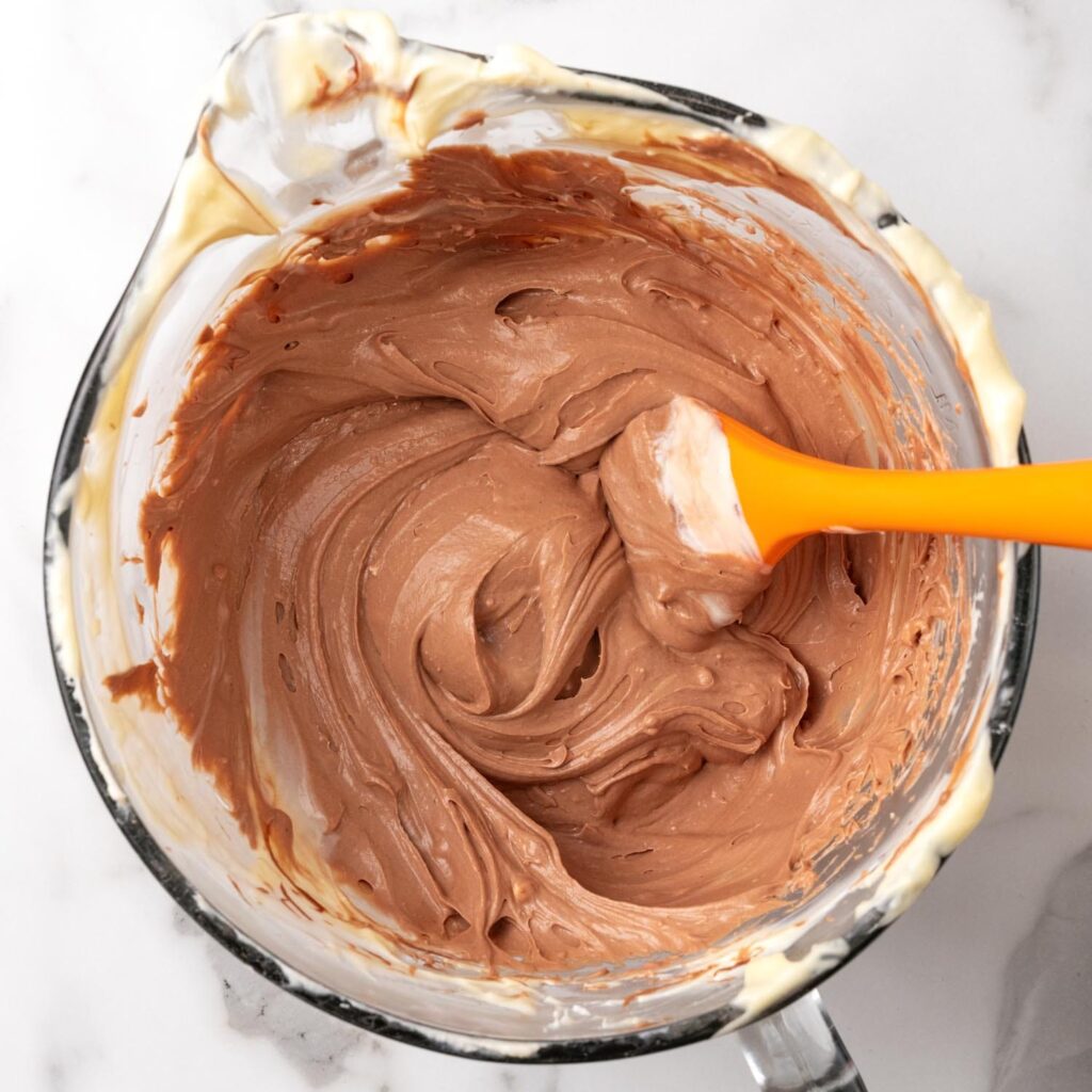 Chocolate cheesecake filling in a glass mixing bowl with an orange spatula.