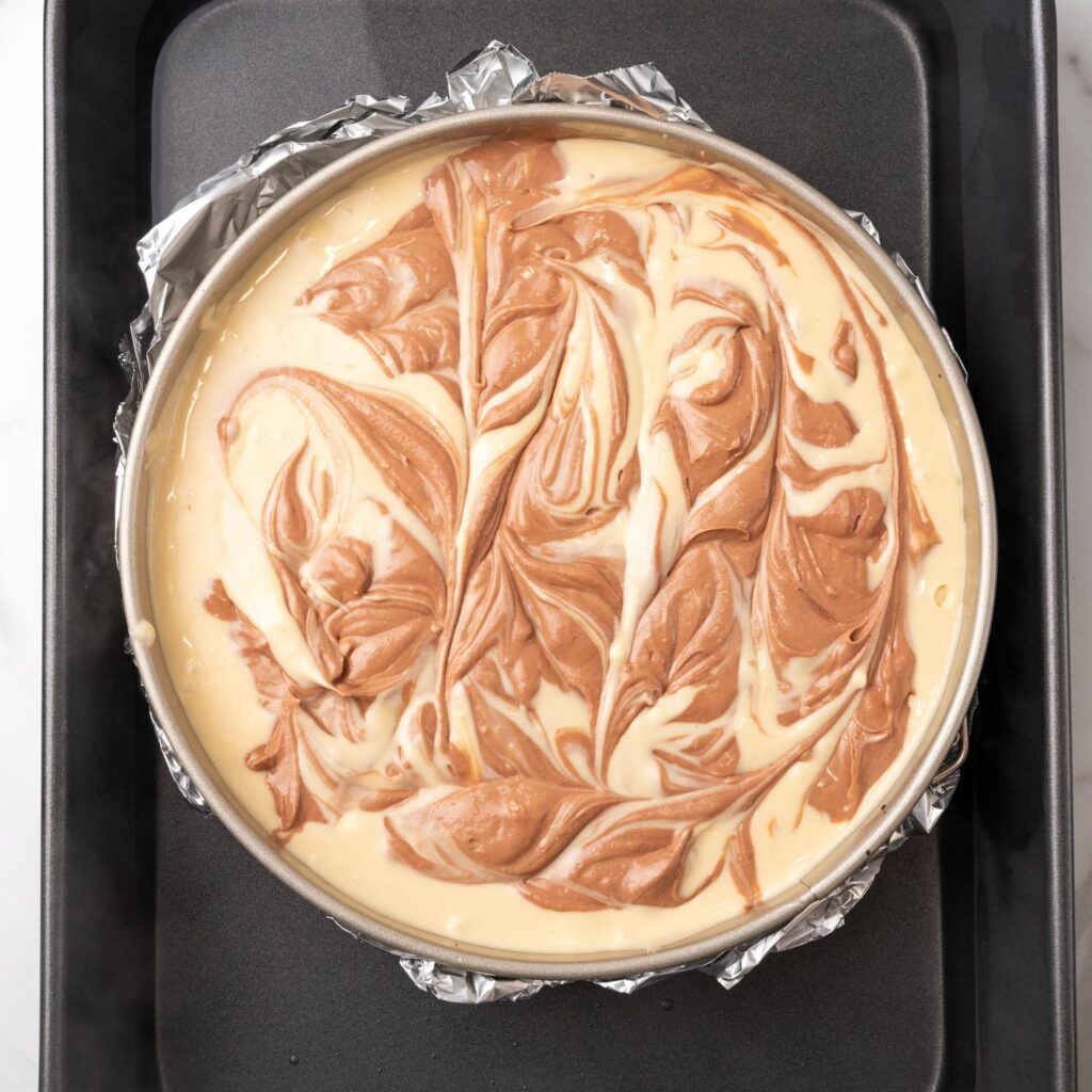 a swirled turtle cheesecake in a spingform pan, wrapped in foil. The pan is placed in a black roaster to create a water bath.