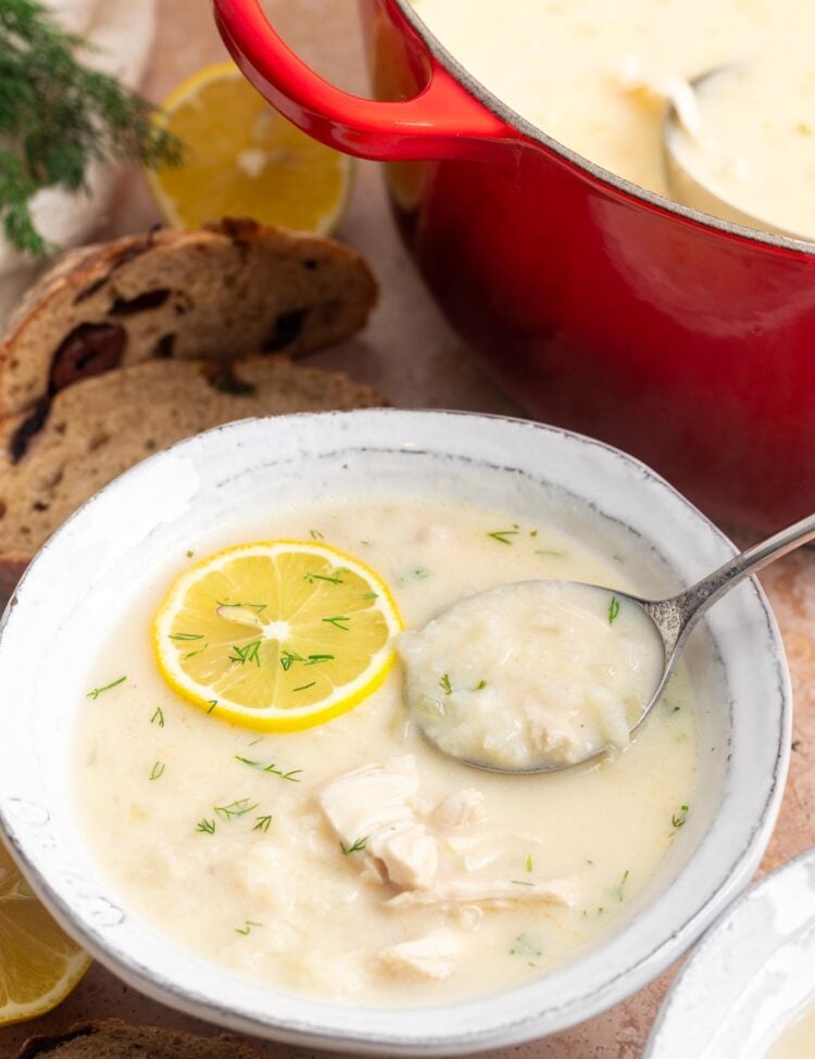 a rustic ceramic bowl of greek lemon chicken soup next to a red pot of the same. The soup is garnished with dill and a lemon slice.