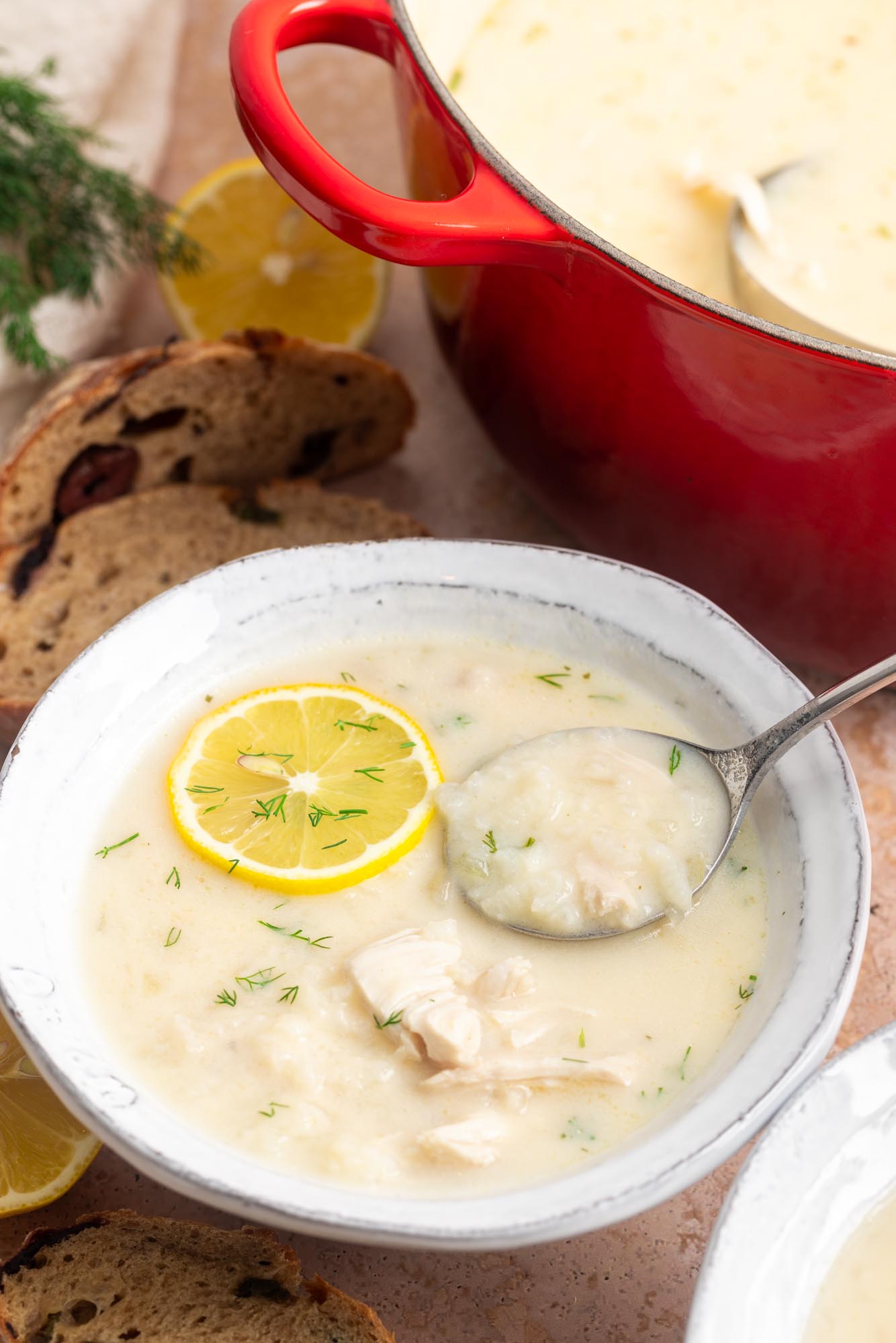 a rustic ceramic bowl of greek lemon chicken soup next to a red pot of the same. The soup is garnished with dill and a lemon slice.