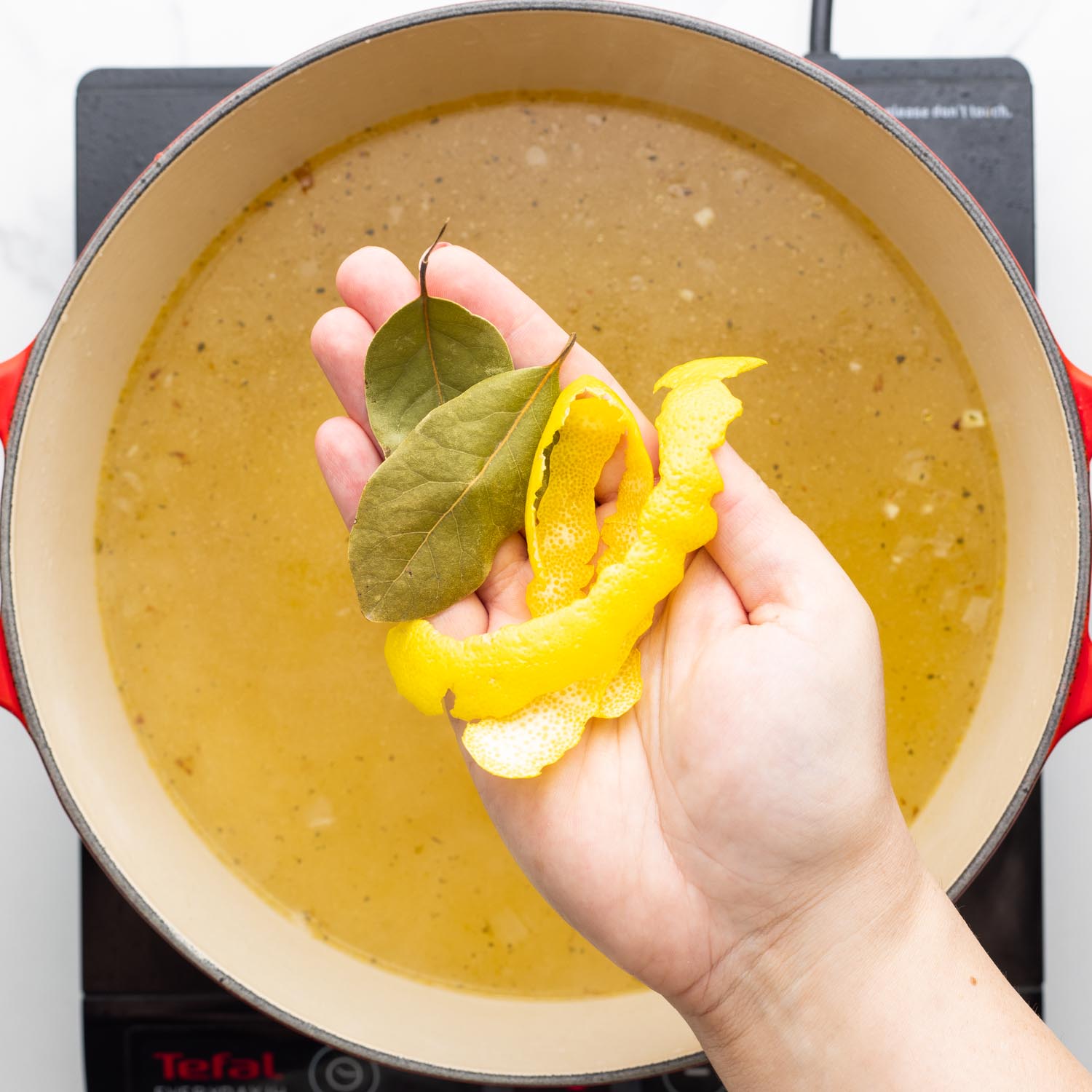 bay leaves and lemon peels held in a hand, over a pot of chicken stock.